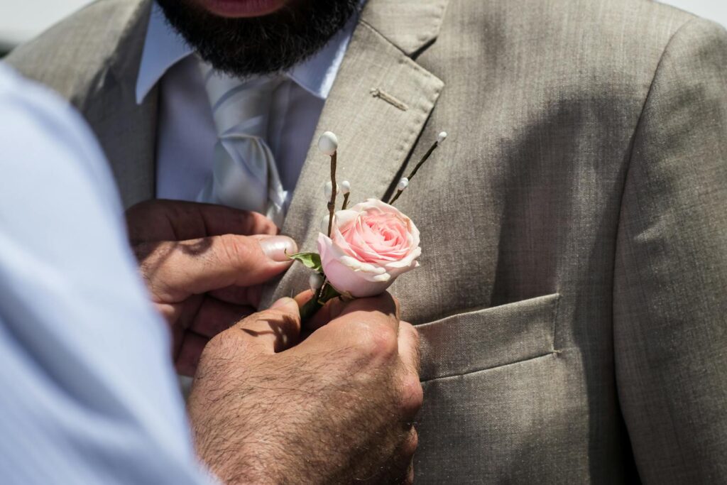 A close-up of a man attaching a pink rose boutonniere to a groom's suit at a wedding.