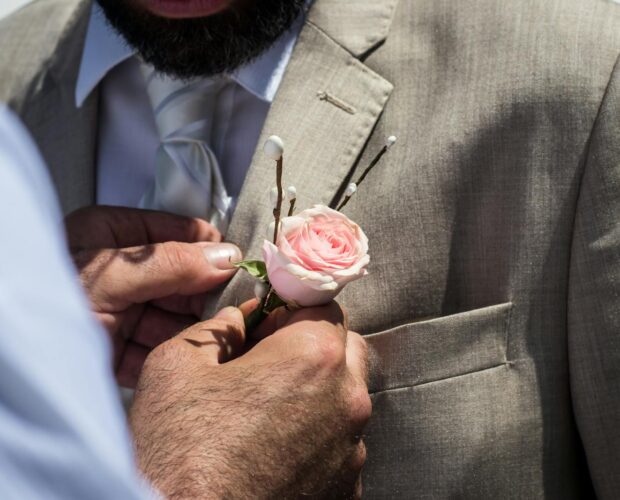 A close-up of a man attaching a pink rose boutonniere to a groom's suit at a wedding.